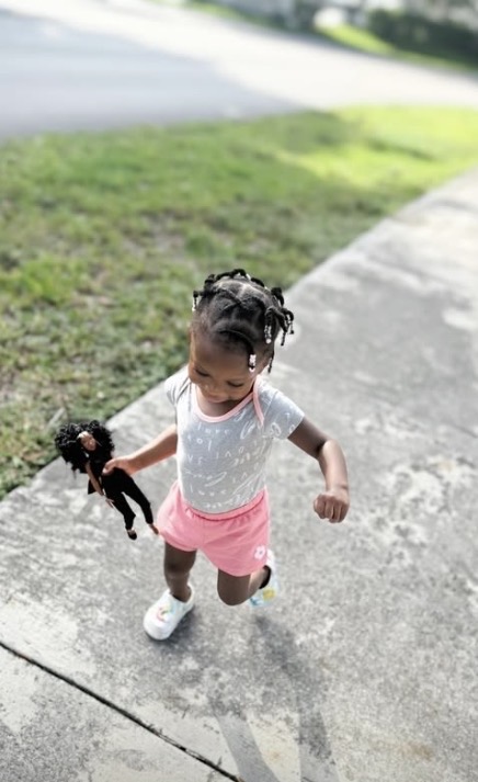 A little girl running down the sidewalk holding her Melanie doll